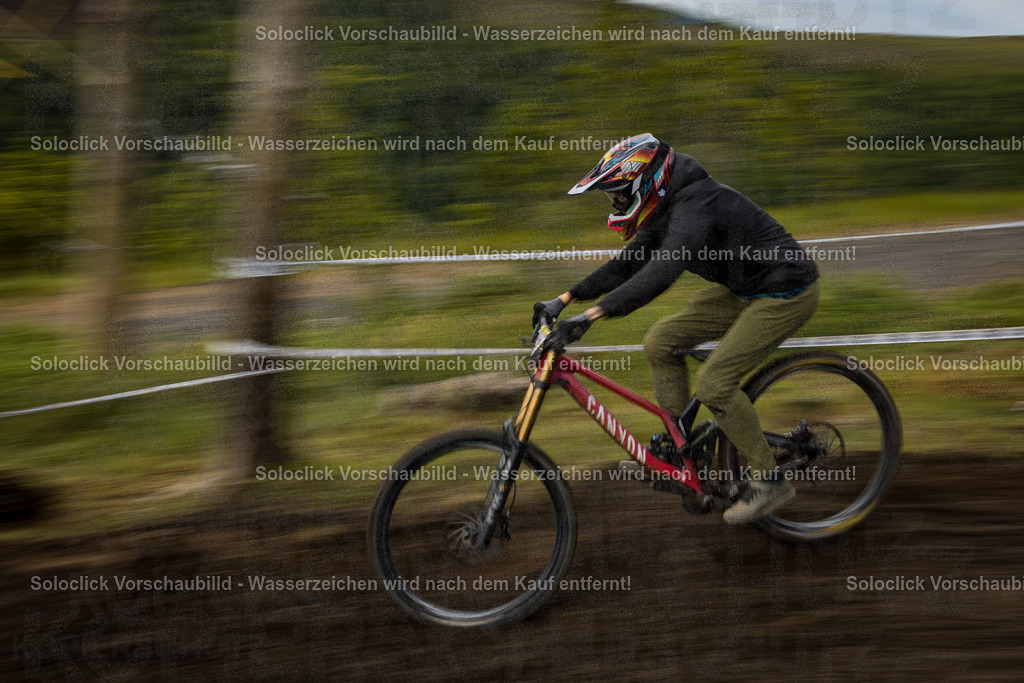 Adrenalin pur: Downhill-Action im Bikepark Winterberg | Ein Mountainbiker, der einen Downhill-Rennen bestreitet, ist im Fokus dieses dynamischen Bildes. Er rast mit hoher Geschwindigkeit einen staubigen Trail hinunter, wobei die Bewegung durch die Unschärfe im Hintergrund und den verschwommenen Vordergrund betont wird. Der Fahrer trägt einen auffälligen Helm und Schutzkleidung, was auf die Intensität des Rennens hinweist. Das Bild fängt die Energie und den Nervenkitzel des Downhill-Mountainbikens ein. - Realisiert mit Pictrs.com