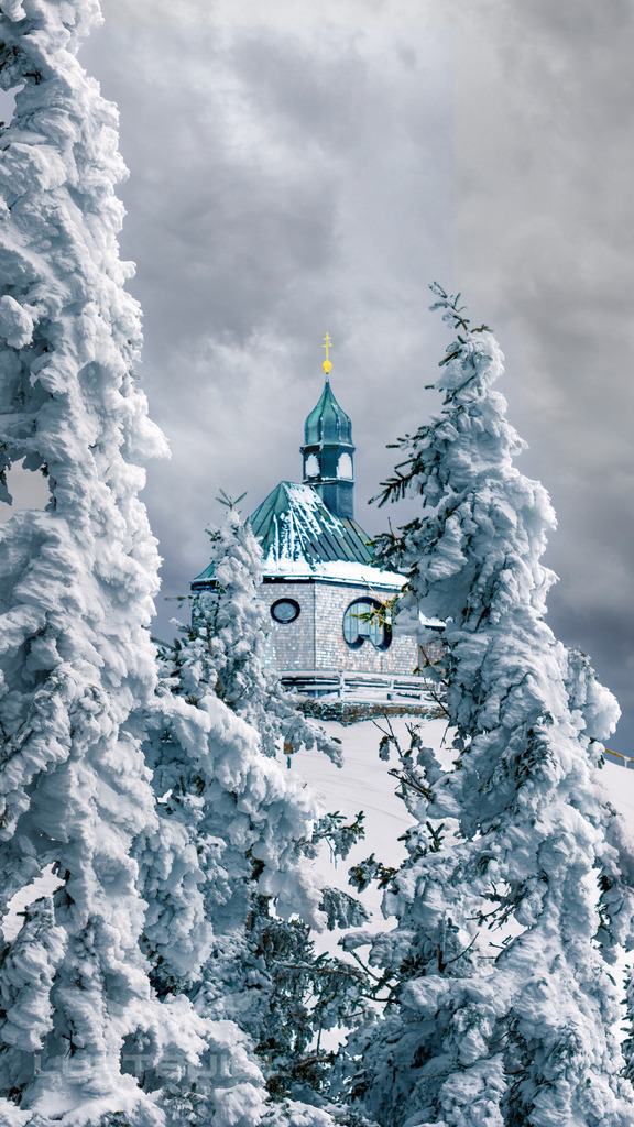 Blick aufs Wallbergkircherl (Perspektivwechsel); W47 | Das Wallbergkircherl zeigt sich hier in einem eindrucksvollen Perspektivwechsel: Eingebettet zwischen schwer verschneiten Bäumen ragt der markante Turm ruhig in den winterlichen Himmel. Die dichte Schneelast auf den Ästen rahmt das Kirchlein beinahe wie ein natürliches Fenster ein und verleiht der Szene eine fast mystische Stimmung – ein stiller, kraftvoller Moment hoch oben am Wallberg. - Realisiert mit Pictrs.com