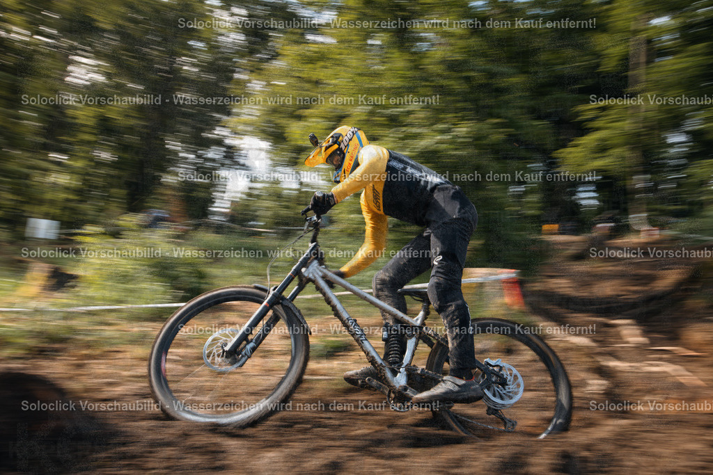 Rasante Fahrt | Ein Mountainbiker in voller Ausrüstung rast auf seinem Downhill-Mountainbike einen staubigen, kurvigen Pfad hinunter. Die Bewegung des Fahrers und des Fahrrads ist durch die lange Belichtungszeit unscharf dargestellt, was die Geschwindigkeit und Dynamik der Szene betont. Der Fahrer trägt einen gelben Helm und eine passende Weste, kombiniert mit schwarzen Hosen. Das Mountainbike ist ein Downhill-Modell mit dicker Federgabel und breiten Reifen. Der Hintergrund besteht aus dichtem Grünwerk und einem schmalen, weißen Zaun. - Realisiert mit Pictrs.com