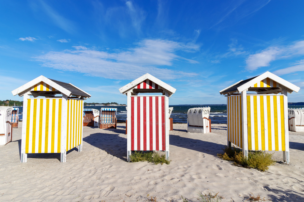 Wandbild: Farben des Sommers – Umkleidekabinen in Eckernförde | Dieses Wandbild zeigt die farbenfrohen Umkleidekabinen am Sandstrand von Eckernförde an der Ostsee. Die Kabinen stehen in klarer Reihe, ihre vertikalen Streifen in Gelb-Weiß und Rot-Weiß leuchten im Sonnenlicht und setzen lebendige Akzente vor dem blauen Himmel. Dahinter reihen sich klassische weiße Strandkörbe entlang des Strandes – nummeriert und ausgerichtet zum Meer. Sie verleihen der Szene eine typische norddeutsche Strandatmosphäre: geordnet, ruhig und einladend. Die Komposition lebt vom Zusammenspiel aus Farbe, Struktur und Küstenkultur – ein Bild, das Sommerstimmung und maritimes Flair vermittelt. Ideal als Wandbild für helle, naturnahe Wohnkonzepte – ob als Leinwandbild, Acrylglasbild, Alu-Dibond FineArt Print oder als Akustikbild. Ein frischer, fröhlicher Akzent für Wohnzimmer, Büro oder Ferienwohnung. - Realisiert mit Pictrs.com