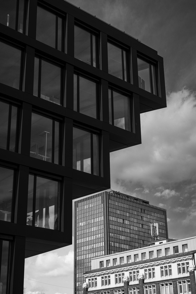 Architektur in Schichten | Diese Schwarzweißaufnahme zeigt das Spannungsfeld zwischen moderner und klassischer Stadtarchitektur in einer klaren, grafischen Komposition. Die markante, auskragende Fassade im Vordergrund dominiert das Bild mit ihrer strengen Formensprache, während sich im Hintergrund weitere architektonische Ebenen zu einem vielschichtigen urbanen Raum verdichten.Licht, Struktur und Perspektive erzeugen eine ruhige, zugleich kraftvolle Bildwirkung, in der sich Massivität und Offenheit gegenüberstehen. Die reduzierte monochrome Darstellung lenkt den Blick auf Geometrie, Rhythmus und architektonische Präsenz und verleiht der Szene eine zeitlose Fine-Art-Ästhetik.Diese Aufnahme übersetzt urbane Architektur in eine moderne Wandarbeit mit klarer grafischer Wirkung – ideal für minimalistische, designorientierte Räume. - Realisiert mit Pictrs.com
