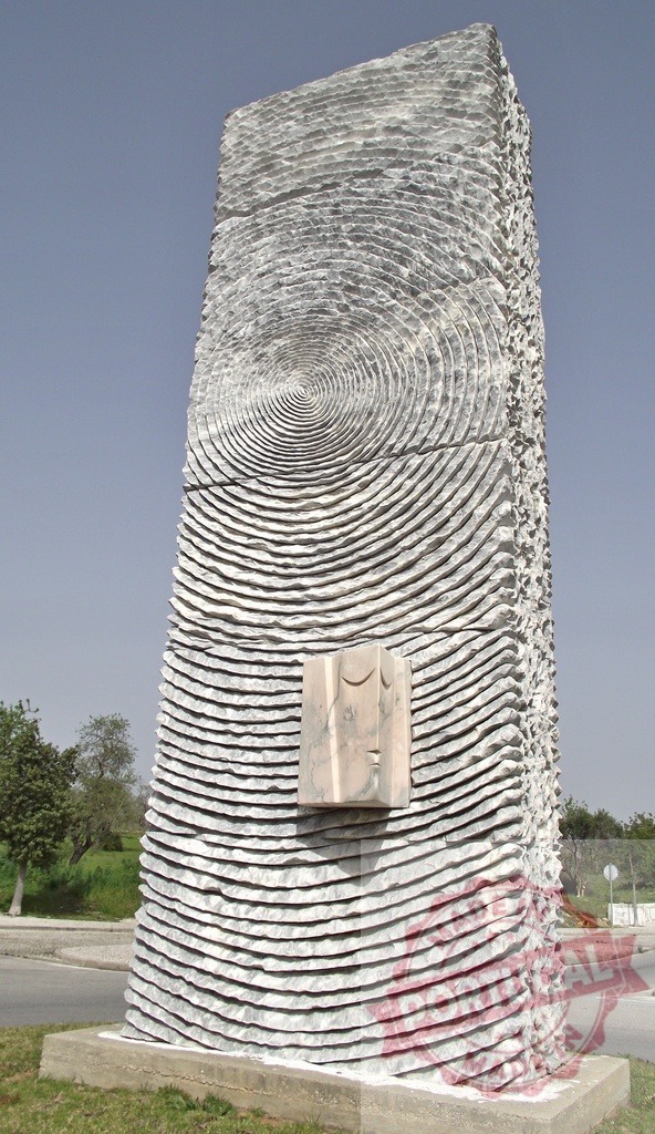 Stone monument pillar with abstract carved circles and mask element. Algarve - Silves | Tall stone pillar with concentric circles and a relief mask standing outdoors - Realized with Pictrs.com