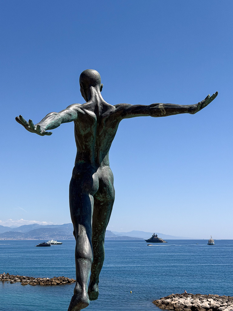 Figure Facing the Sea | The rear view of a contemporary sculpture near the Picasso Museum in Antibes, France. With open arms facing the Mediterranean horizon, the figure creates a poetic atmosphere of freedom, balance and connection between art and sea. ✨ - Realisiert mit Pictrs.com