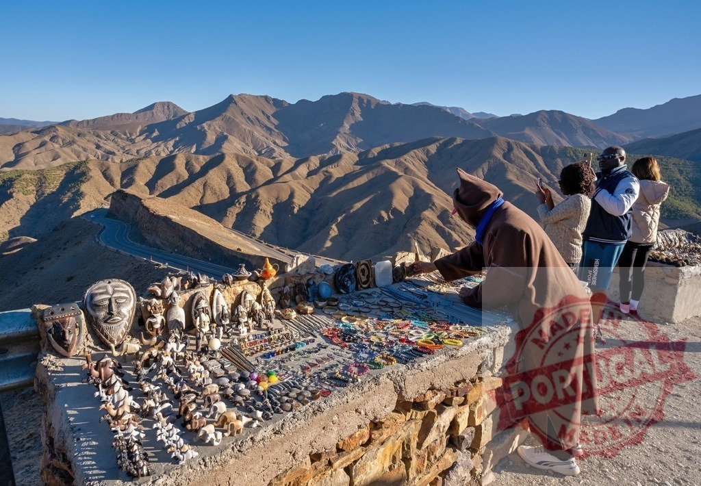 Moroccan vendor arranging souvenirs on stall in High Atlas mountains with tourists taking pictures | Berber vendor arranging his souvenirs on a stall in the Tizi n'Tichka pass in the High Atlas mountains while tourists take pictures of the landscape - Realized with Pictrs.com