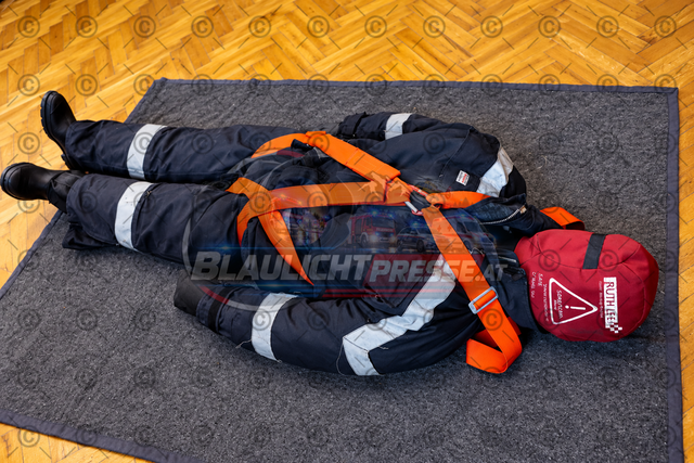 Feuerwehrübung mit Feuerwehrdummy | Ein Übungsdummy in Feuerwehrschutzkleidung liegt gesichert auf einer Übungsmatte. Mithilfe eines orangefarbenen Rettungsgurts können verschiedene Rettungstechniken und der sichere Transport verletzter Personen bei Feuerwehreinsätzen trainiert werden. - Realisiert mit Pictrs.com