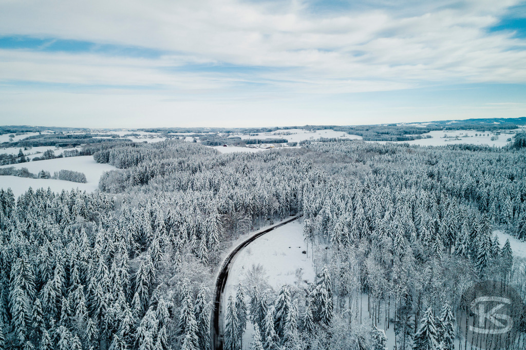 Winterlandschaft Oberschwaben – Verschneiter Wald und Straße aus der Luft | Diese Drohnenaufnahme zeigt eine weitläufige Winterlandschaft in Oberschwaben. Eine geschwungene Straße führt durch dichten, schneebedeckten Nadelwald und öffnet sich zu verschneiten Feldern. Die Luftperspektive offenbart die sanften Hügel und die klare Struktur der Landschaft unter weitem Winterhimmel. - Realisiert mit Pictrs.com
