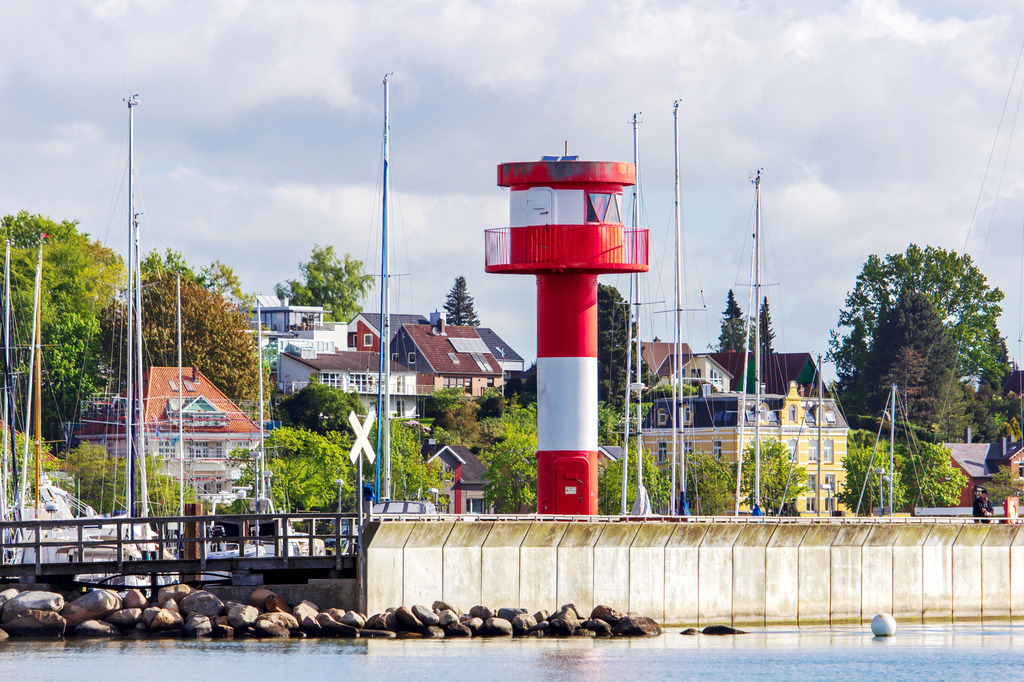 Wandbild: Leuchtturm in Eckernförde am Yachthafen | Dieses Wandbild im Querformat zeigt den malerischen Leuchtturm am Yachthafen in Eckernförde. Im Hintergrund sind die Masten der Segelboote sowie die einige Häuser zu sehen. - Realisiert mit Pictrs.com