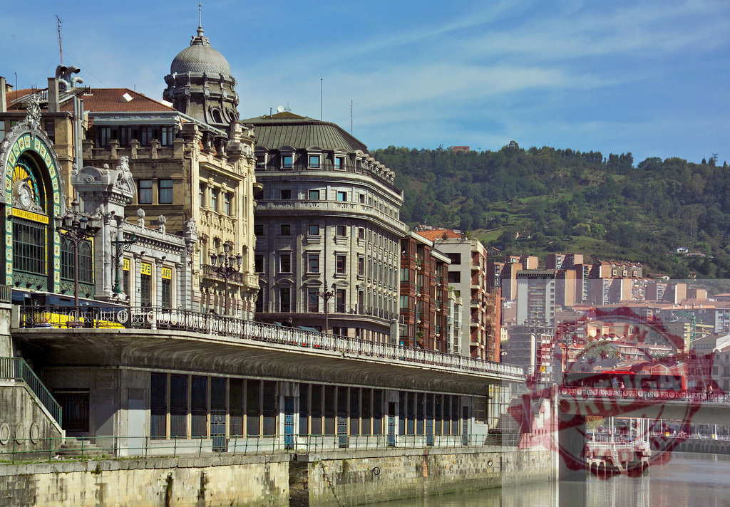 Bilbao city view with Estacion de la Concordia building and Nervion River. Spain - Bilbao | Bilbao cityscape featuring historic architecture, the Nervion River, and green hills under a blue sky - Realized with Pictrs.com