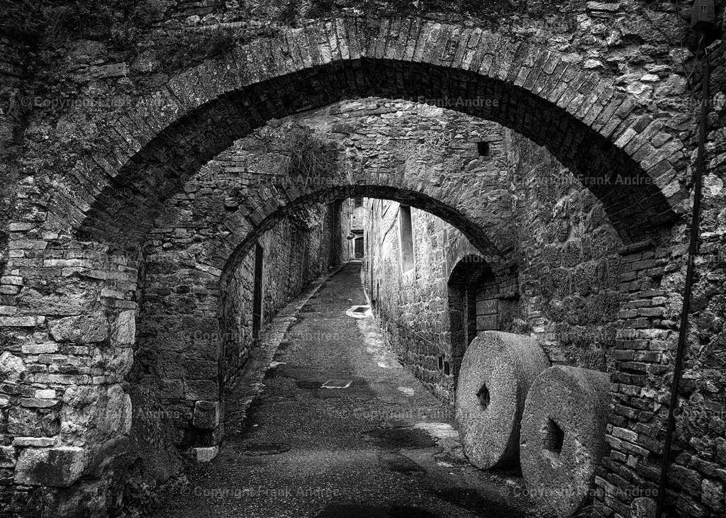 San Gimignano | Blick in eine enge Gasse mit Torbögen und alten Mühlrädern in der Altstadt von San Gimignano. Schwarz weiß Fotografie aus der Toskana. - Realisiert mit Pictrs.com