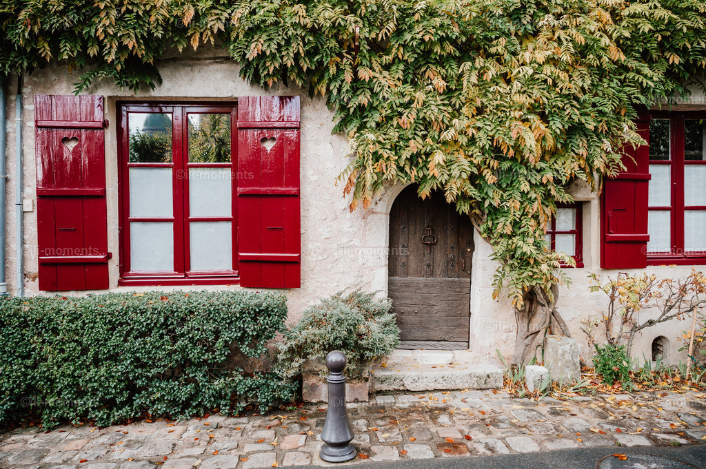 die Märchentür | „Die Märchentür“ ist ein bezauberndes Fotomotiv, das die Magie eines alten französischen Sandsteinhauses in der herbstlichen Jahreszeit einfängt. Im Mittelpunkt steht eine kleine, alte Holztür mit Bogen, die fast hinter den üppigen Blättern des Blauregens verschwindet. Schon die ersten Herbstfarben zeigen sich an den Blättern und verleihen der Szenerie eine warme, goldene Atmosphäre. Die knallroten Fensterläden und Fensterrahmen setzen einen lebendigen Kontrast zum Sandstein und dem herbstlich getönten Grün der Pflanzen.Dieses Bild verbindet romantischen Dorfcharme mit herbstlicher Stimmung und eignet sich ideal für Wohnzimmer, Flure, Küchen, Cafés oder Boutique-Hotels, die eine warme, verträumte Atmosphäre suchen. - Realisiert mit Pictrs.com