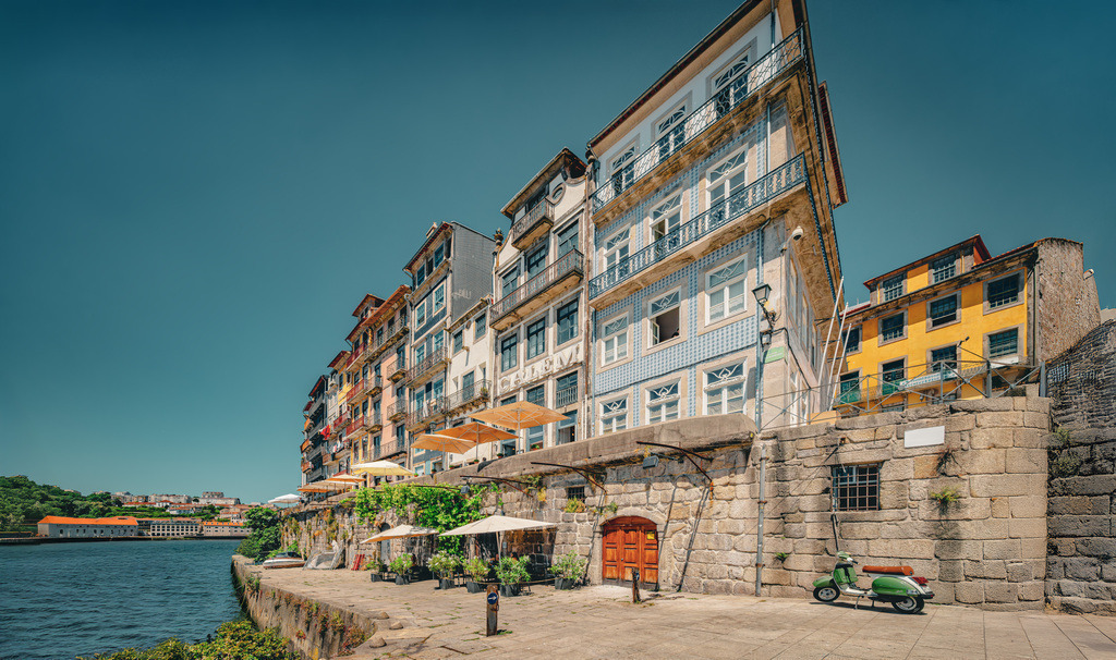 Eine Uferszene am Douro in Porto, Schirme, ein Motorroller | Die Szene am Ufer des Douro zeigt die Cais da Ribeira, die berühmte Uferpromenade im historischen Ribeira-Viertel von Porto, Portugal. Dieses Viertel ist das Herzstück der Stadt und gehört zum UNESCO-Weltkulturerbe. Details zur Ribeira-UferpromenadeLage: Die Promenade erstreckt sich entlang des Nordufers des Douro-Flusses in der Altstadt von Porto.Atmosphäre: Sie ist bekannt für ihre mittelalterlich anmutenden Gassen, lebhaften Bars und Restaurants.Sehenswürdigkeiten: Die Uferpromenade bietet einen malerischen Blick auf die traditionellen, bunten Häuser, die den Hang hinaufführen, und auf die gegenüberliegende Stadt Vila Nova de Gaia. Im Hintergrund sind die Türme der Kathedrale (Sé Catedral) sichtbar. - Realisiert mit Pictrs.com