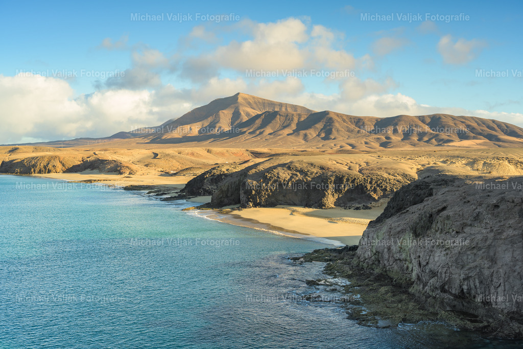 Playa de Papagayo und Hacha Grande auf Lanzarote | Am Playa de Papagayo trifft die weiche Linie der goldenen Bucht auf die markante Silhouette des Hacha Grande, der über dem Süden Lanzarotes wie ein ruhiger, dunkler Ankerpunkt steht. Vom Strand aus wirkt das Massiv fast skulptural und verleiht der Szenerie eine besondere Tiefe. - Realisiert mit Pictrs.com