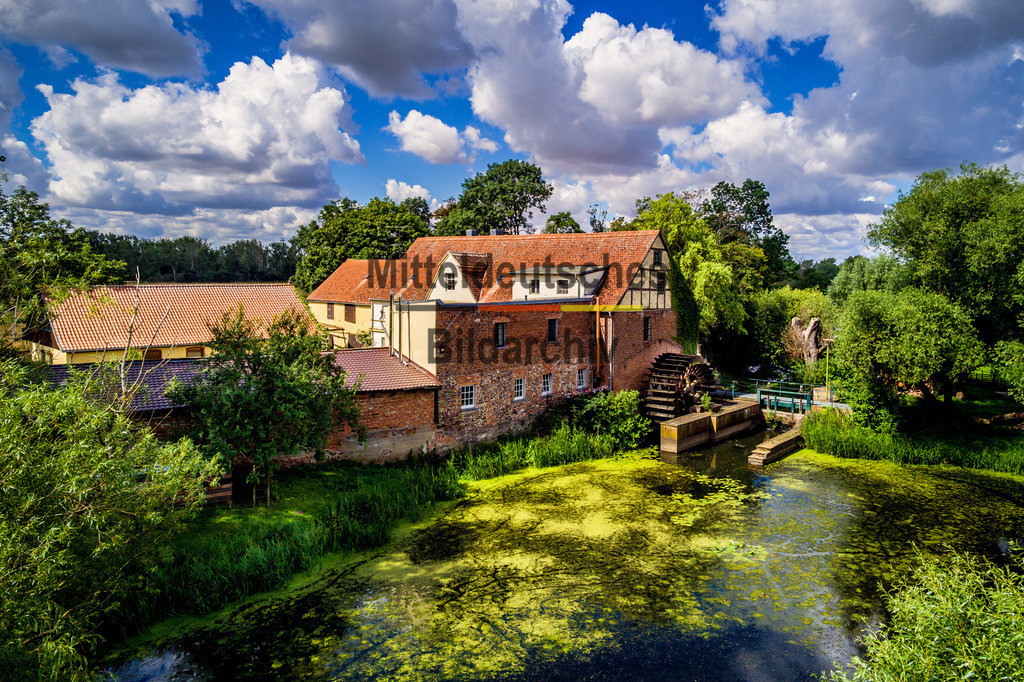 Wassermühle Elbeu bei Wolmirstedt-0005 | Das Luftbild zeigt die historische Mühle in Elbeu aus einer Drohnenperspektive an einem sonnigen Sommertag. Im Vordergrund liegt eine ruhige Wasserfläche, während über der Mühle markante Quellwolken den Himmel prägen und der Aufnahme eine eindrucksvolle Tiefe verleihen. Die farbenfrohe Landschaft bildet einen harmonischen Rahmen für das technische Denkmal.Die Mühle Elbeu steht beispielhaft für die Entwicklung der Getreideverarbeitung vom handwerklichen Mühlenbetrieb zur industriellen Produktion. Im Laufe ihrer Geschichte wurde die Antriebstechnik mehrfach modernisiert – von der ursprünglichen Wind- beziehungsweise Wasserkraft bis hin zum elektrischen Antrieb. Walzenstühle, Elevatoren und Förderanlagen ersetzten nach und nach die traditionellen Mahlgänge und steigerten die Leistungsfähigkeit der Mühle erheblich. Solche Mühlen konnten täglich mehrere Tonnen Getreide zu Mehl und Futtermitteln verarbeiten und waren über Jahrzehnte ein wichtiger Bestandteil der regionalen Landwirtschaft.Die Drohnenaufnahme verbindet historische Mühlentechnik mit der sommerlichen Landschaft der Elbaue. Die markanten Quellwolken, das Wasser im Vordergrund und die eindrucksvolle Architektur der Mühle machen das Bild zu einer gelungenen Verbindung aus Technik, Landschaft und Industriegeschichte. - Realisiert mit Pictrs.com