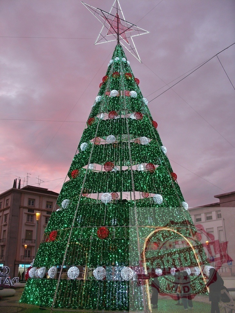 Giant illuminated christmas tree standing tall in city square | Huge christmas tree decorated with lights and ornaments brightening a city square at dusk - Realized with Pictrs.com