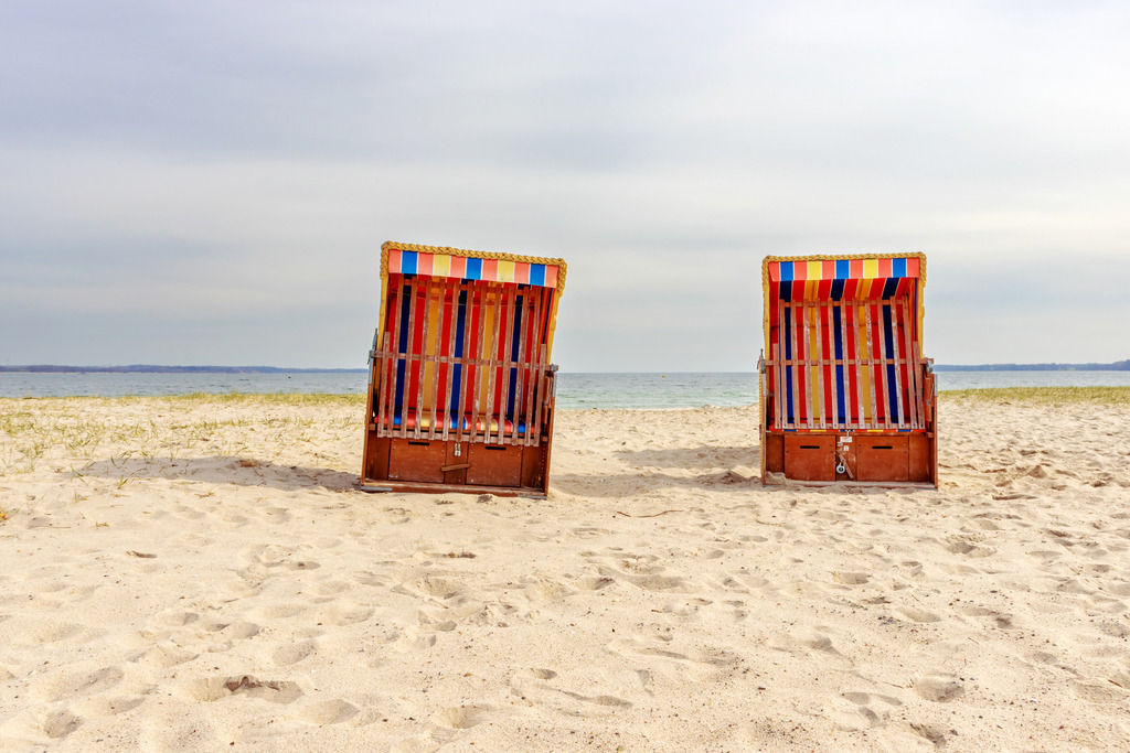 Wandbild: Strandkörbe am Strand in Eckernförde | Dieses Wandbild im Querformat zeigt zwei Strandkörbe am Strand in Eckernförde. Der Himmel ist größtenteils bewölkt, dennoch scheint die Sonne auf den Strand.  - Realisiert mit Pictrs.com