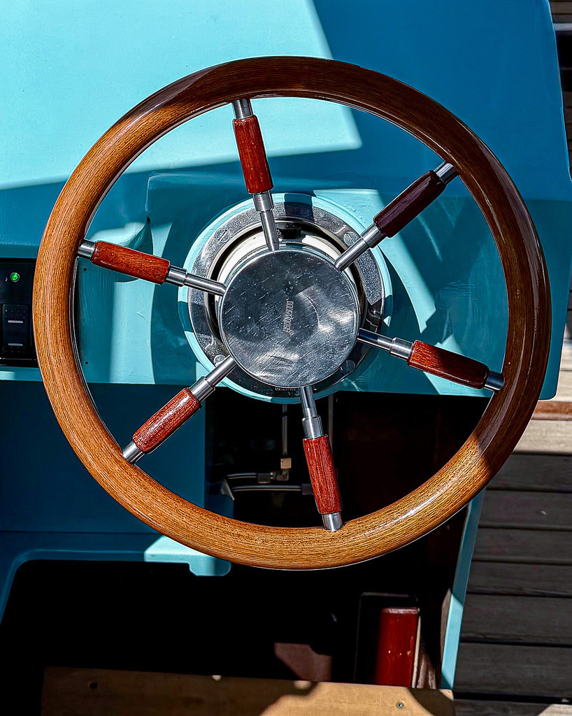 Maritimes Steuerrad – Minimalistisches Schiffs Wandbild aus Holz | Exklusive und detailreiche maritime Fotokunst: Dieses High-End Wandbild zeigt eine minimalistische Nahaufnahme eines klassischen Schiffslenkrads vor einer hellblauen Schiffswand. Der faszinierende Kontrast aus warmen, polierten Holzstrukturen und dem maritimen Hintergrund bringt frisches Küsten-Feeling und zeitlose Eleganz in Museumsqualität an deine Wände. Ein perfekter Blickfang auf Alu-Dibond oder Acrylglas für Badezimmer, Ferienwohnungen oder moderne Wohnkonzepte. - Realisiert mit Pictrs.com