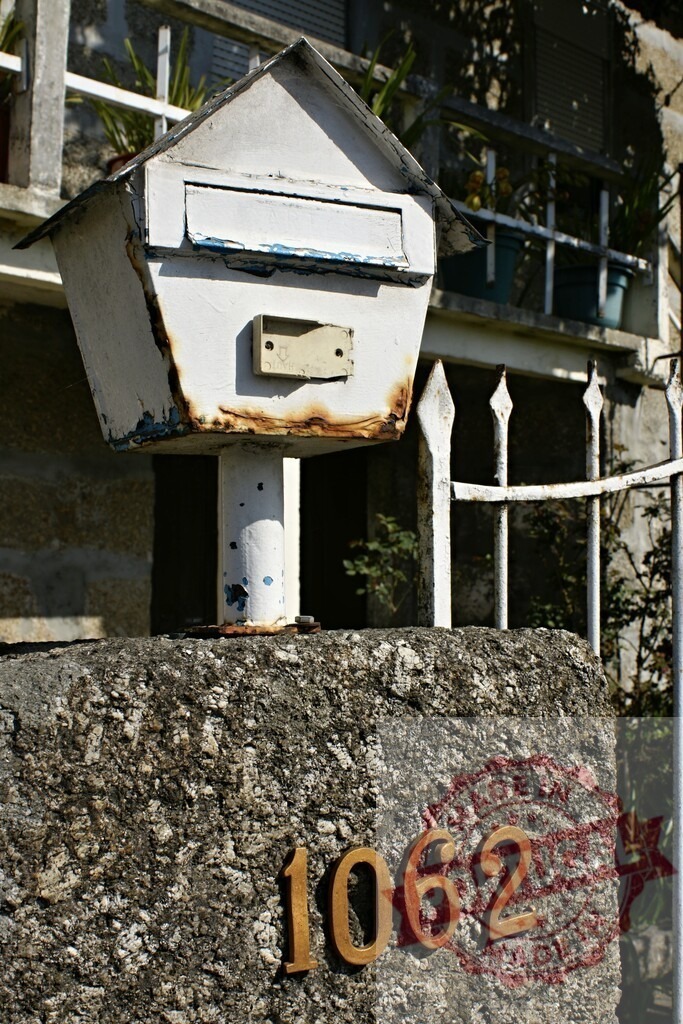 Old rusty mailbox standing on a stone pillar with house number 1062 | Old rusty mailbox standing on a stone pillar with the house number one thousand and sixty-two in front of a house with white fence - Realized with Pictrs.com