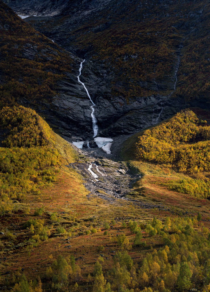 Wasserfall im Tal Austerdalen (Norwegen) im Herbst 2021 | Im Schatten einer engen Bergkluft, schlängelt sich ein Wasserfall zu Tal. Das Licht der Sonne streift den dichten Bewuchs der Berghänge, während in den ganzjährig schattigen Bereichen noch Schneereste des letzten Winters liegen geblieben sind. - Realisiert mit Pictrs.com