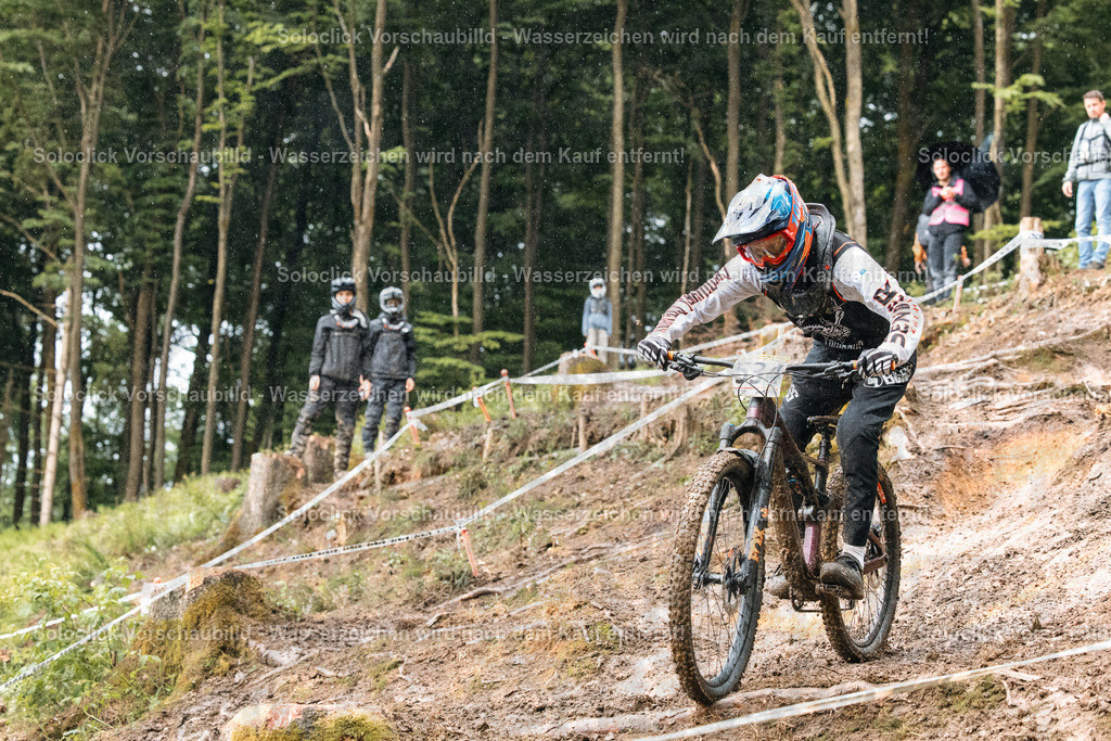 Mountainbike-Action im Gelände | Ein Mountainbiker rast einen steilen, unebenen Pfad hinunter, umgeben von dichtem Grün und Bäumen. Der Fahrer trägt einen auffälligen Helm und Schutzkleidung und konzentriert sich voll und ganz auf die Fahrt. Zuschauer stehen am Wegesrand und beobachten das Rennen. Der Pfad ist mit Bändern markiert, was auf einen Wettkampf hindeutet. Der Himmel ist bewölkt, was auf feuchte Bedingungen hindeutet. - Realisiert mit Pictrs.com