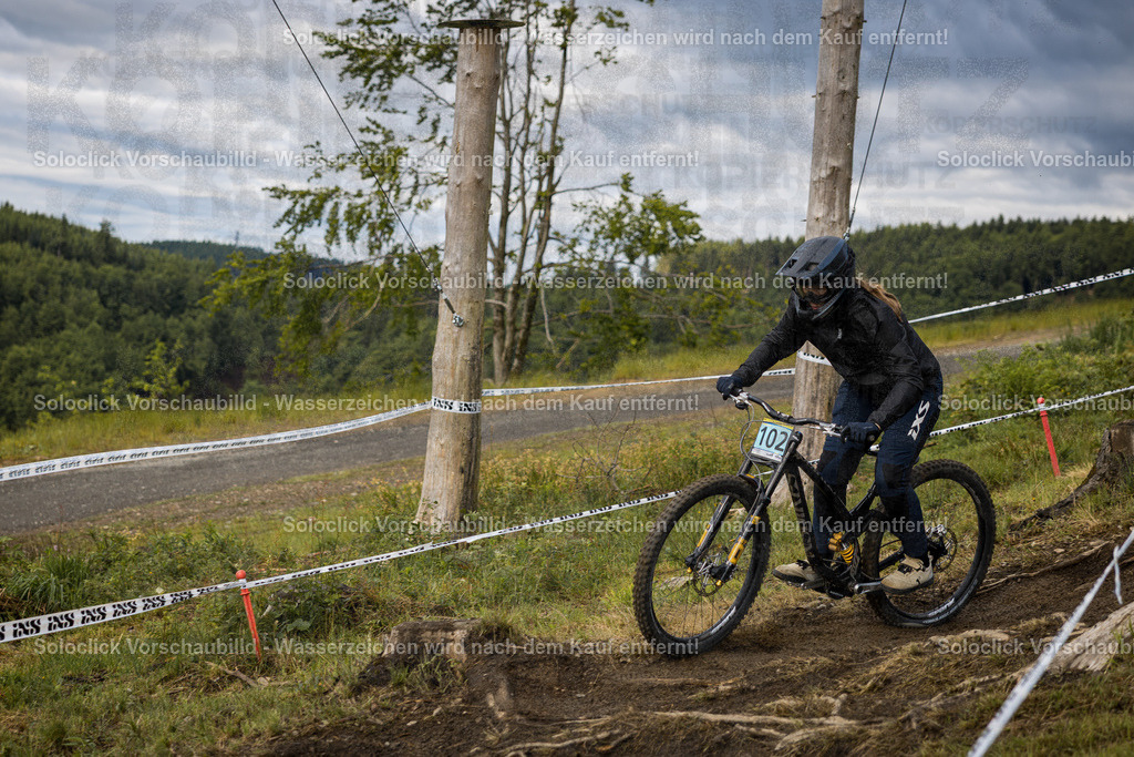 Action im Bikepark Winterberg | Ein Mountainbiker stürzt sich mit hoher Geschwindigkeit einen Downhill-Kurs hinunter. Er trägt einen schwarzen Helm und eine schwarze Jacke und fährt ein gelb-schwarzes Mountainbike mit der Nummer 102. Der Kurs ist mit roten und weißen Bändern markiert, und im Hintergrund erstreckt sich ein dichter Wald. - Realisiert mit Pictrs.com