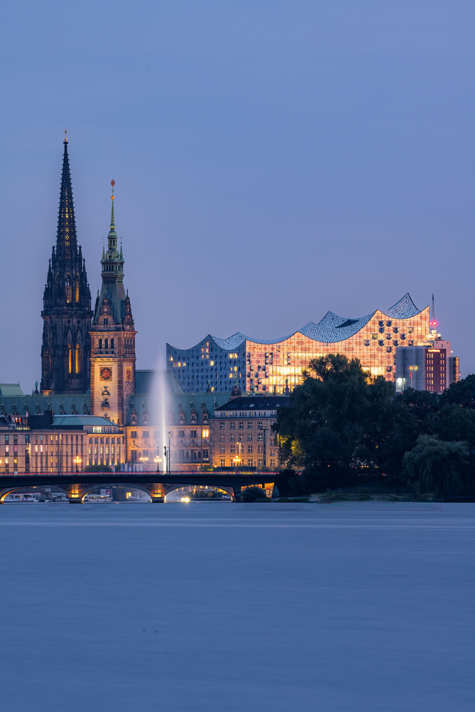 Hamburger Innenstadt an der Alster zur blauen Stunde | Blick über das ruhige Wasser auf die Hamburger Innenstadt zur blauen Stunde. Die beleuchtete Uferpromenade, markante Kirchtürme und die abendliche Skyline verleihen der Aufnahme eine ruhige und elegante Stimmung. - Realisiert mit Pictrs.com