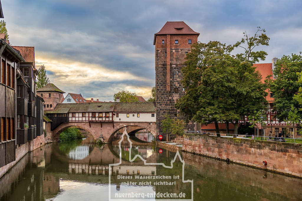 Nürnberger Stadtblick, 05.10.24 | Das Henkerhaus in Nürnberg, das direkt über der Pegnitz erbaut ist, neben dem historischen Wasserturm, der heute als Studentenwohnheim dient. Die Fachwerkarchitektur des Henkerhauses und der massive Steinbau des Turms spiegeln sich im ruhigen Wasser wider. Die Szene ist von Bäumen und dem gemauerten Ufer gesäumt und wird durch die untergehende Sonne beleuchtet. - Realisiert mit Pictrs.com