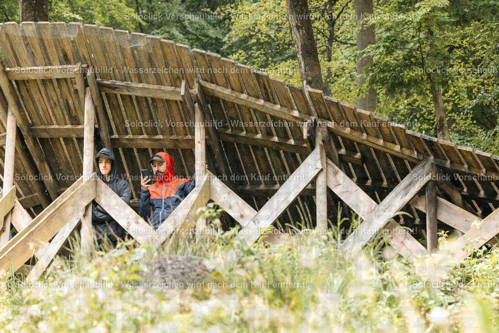 Versteckte Momente im Wald | Das Bild zeigt zwei Personen, die hinter einem Holzzaun in einem Wald stehen. Eine der Personen hält ein Smartphone und scheint zu fotografieren oder ein Video aufzunehmen. Der Zaun besteht aus Holzlatten und -balken und ist teilweise verfallen. Das Gras um den Zaun ist hoch und grün. Im Hintergrund sind weitere Bäume und Vegetation zu sehen. - Realisiert mit Pictrs.com