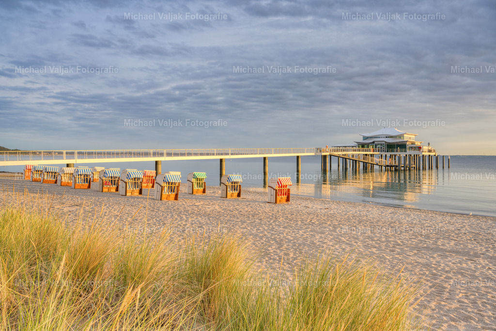 Morgens am Timmendorfer Strand | An einem bewölkten Morgen schafft es die Sonne kurz durch die Wolkendecke und taucht die Seeschlösschenbrücke mit dem Teehaus am Ende und die Strandkörbe sowie das Dünengras im Vordergrund in ein warmes Licht.  - Realisiert mit Pictrs.com