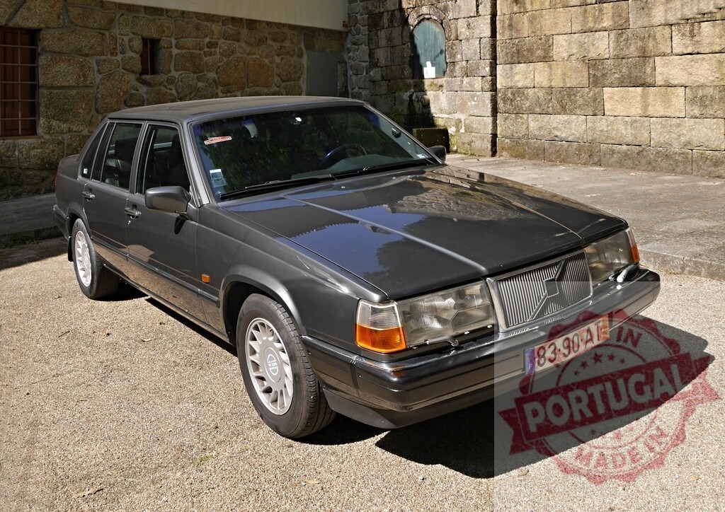 Volvo 940 sedan parking in front of stone wall | Gray Volvo 940 sedan parked on gravel in front of a rustic stone building - Realized with Pictrs.com