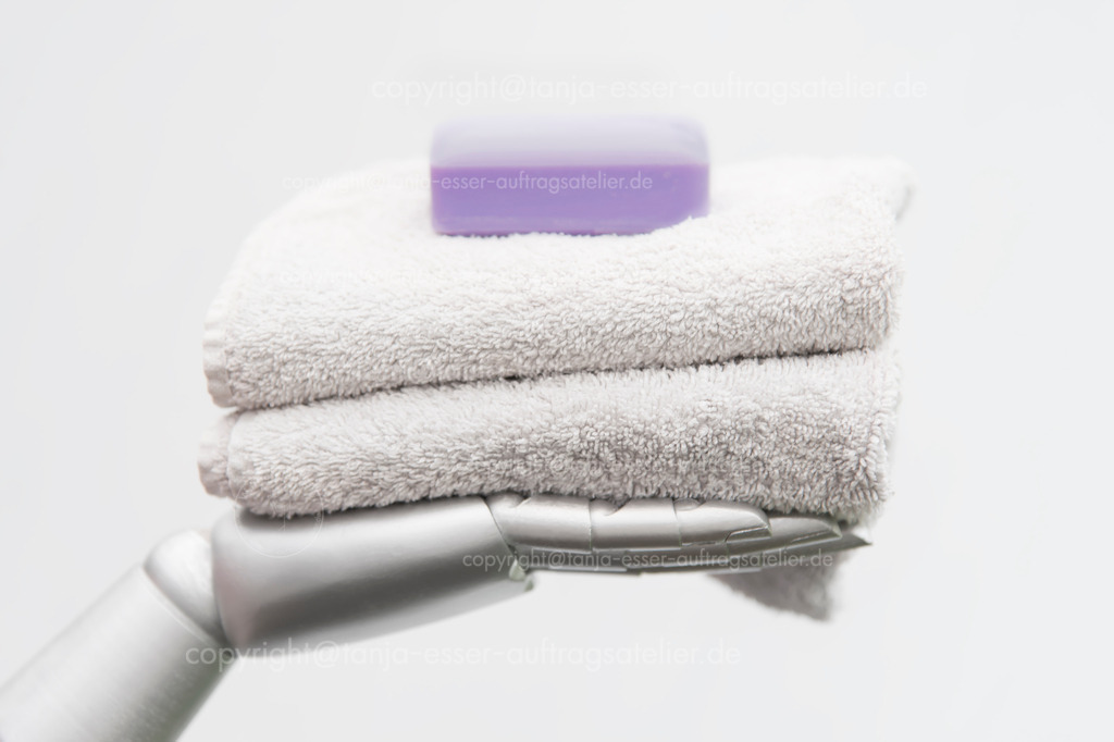 Robot takes care of people | Roboterarm hält Frotteehandtücher und ein Stück Seife in der Hand. Das Bild symbolisiert die Pflege von Menschen durch Maschinen. Konzeptuelles Foto vor weißem Hintergrund mit Copy Space.