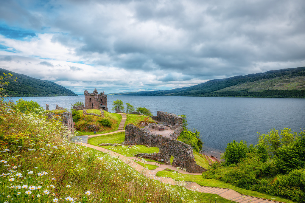 Die malerische Ruine des Urquhart Castle bei Loch Ness,Margeriten im Vordergrund,Schottische Highlands | Das Bild zeigt die malerische Ruine des Urquhart Castle am Ufer des berühmten Loch Ness in den schottischen Highlands.Am Ufer des Loch Ness, nahe dem Ort Drumnadrochit in Schottland.Die Festung stammt in ihren Ursprüngen aus dem 13. Jahrhundert und spielte eine wichtige Rolle in den schottischen Unabhängigkeitskriegen.Die Anlage ist eine der meistbesuchten historischen Sehenswürdigkeiten Schottlands und bietet einen hervorragenden Blick über den See - Realisiert mit Pictrs.com