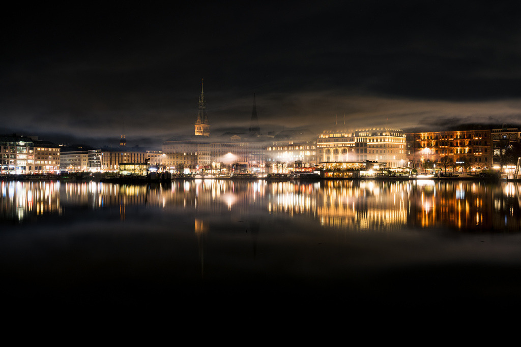 Hamburg Skyline an der Alster bei Nacht | Eine ruhige Panoramaansicht der Hamburger Innenstadt entlang der nächtlichen Alster. Die warmen Lichter der Stadt bilden einen starken Kontrast zum dunklen Himmel und Wasser. - Realisiert mit Pictrs.com