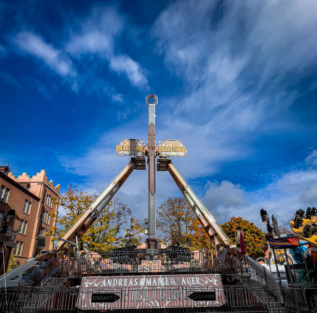 Basel Herbstmesse – Fahrgeschäft Bayern Breaker bei Tag | Spektakuläre Action- und Jahrmarktfotografie aus Basel: Dieses High-End Wandbild fängt den gigantischen Fahrgeschäft-Riesen Bayern Breaker auf der Basler Herbstmesse ein. Vor einem dramatisch blauen Wolkenhimmel entfaltet die mechanische Symmetrie des Karussells ihre volle visuelle Wucht. Ein lebendiges, kraftvolles Kunstwerk voller Adrenalin und Dynamik. Perfekt als moderner Eyecatcher auf Alu-Dibond oder unter edlem Acrylglas. - Realisiert mit Pictrs.com