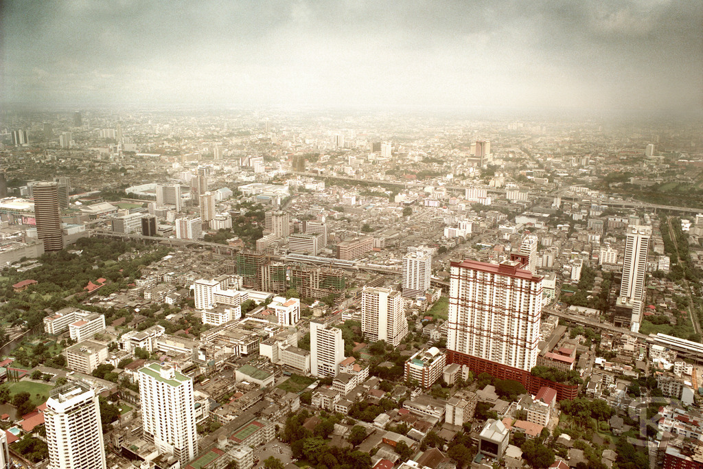Luftbild von Bangkok (2006) mit massivem Autobahnkreuz, aufgenommen vom Baiyoke Tower |  Eine weitläufige Luftaufnahme von der Aussichtsplattform des Baiyoke Sky Tower in Bangkok (Thailand) aus dem Jahr 2006, die das geschäftige Stadtbild und einen komplexen, vielspurigen Autobahnknotenpunkt bei Tag zeigt. Die Aufnahme fängt die dichte urbane Entwicklung und das weitläufige Verkehrsnetz der Metropole ein. - Realisiert mit Pictrs.com
