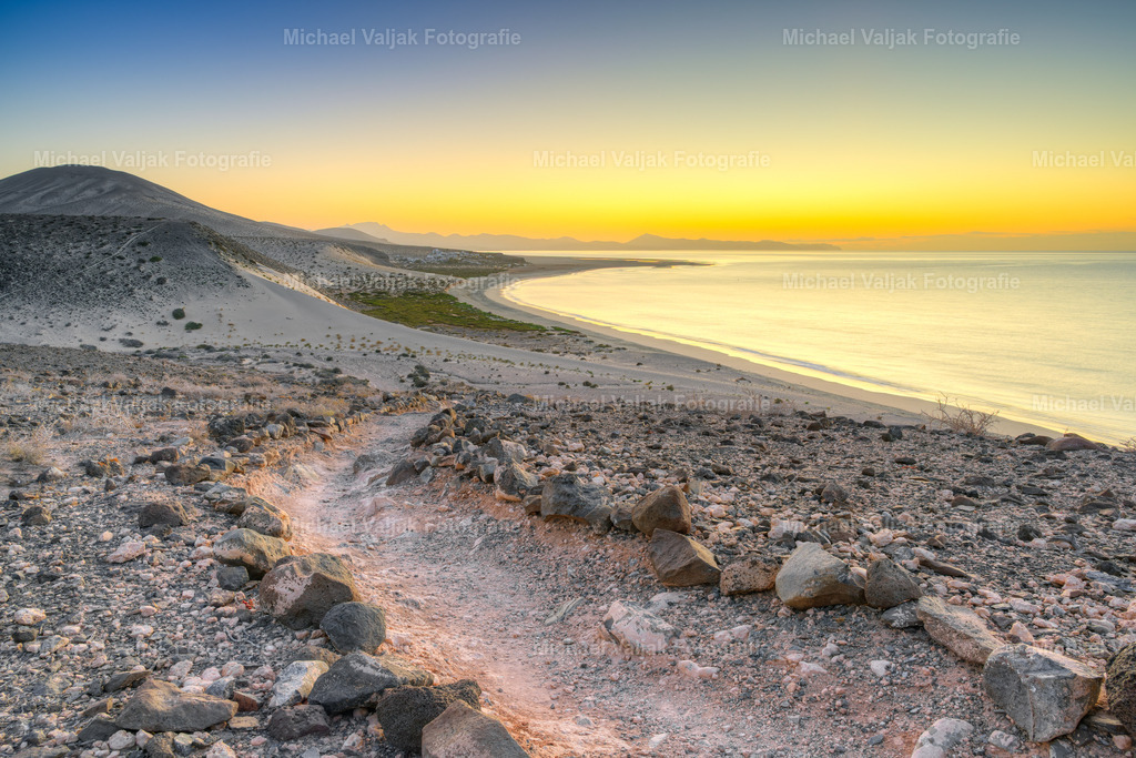 Blick Richtung Playa de Sotavento auf Fuerteventura | In der Morgendämmerung zeigt sich der Blick in Richtung Playa de Sotavento von seiner sanften, fast stillen Seite. Das erste Licht des Tages legt einen zarten Schimmer über die weiten Sandflächen, während die Konturen der Dünen und Lagunen langsam klarer werden. Die Farben sind noch gedämpft und die Landschaft wirkt weit, ruhig und unberührt. Ein Moment, in dem die Insel erwacht und Sotavento seine stille Eleganz zeigt, bevor das kräftige Tageslicht die Szene übernimmt. - Realisiert mit Pictrs.com
