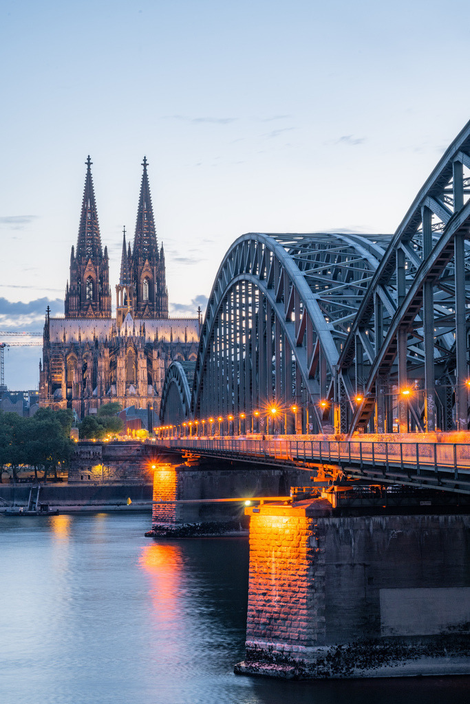 Kölner Dom und Hohenzollernbrücke zur blauen Stunde | Der beleuchtete Kölner Dom erhebt sich hinter der Hohenzollernbrücke, während sich die warmen Lichter im Rhein spiegeln. Das Hochformat betont die markanten Türme und die geschwungene Stahlkonstruktion. - Realisiert mit Pictrs.com