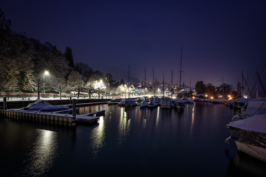Blaues Innehalten | Nächtliche Szene eines kleinen Bootshafens im Winter. Im Vordergrund ruht dunkles, fast schwarzes Wasser, in dem sich die Lichter der Uferpromenade in langen, gelblichen Schlieren spiegeln. Zahlreiche Segel- und Motorboote liegen dicht aneinander an den Stegen; ihre Verdecke sind mit einer dünnen, weissen Schneeschicht bedeckt. Links ragt ein hölzerner Bootssteg ins Wasser, ebenfalls von Schnee überzuckert.Das Ufer wird von hellen Strassenlaternen beleuchtet, die die verschneiten Bäume und die angrenzende Strasse in ein kaltes, weiss-gelbes Licht tauchen. Im Hintergrund ziehen sich sanfte Hügel nach oben, an denen vereinzelt beleuchtete Häuser einer Ortschaft zu erkennen sind. Der Himmel ist tiefdunkelblau bis violett und wirkt durch eine leichte Trübung oder Dunstschicht fast flächig. Die gesamte Atmosphäre ist still, frostig und friedlich. - Realisiert mit Pictrs.com