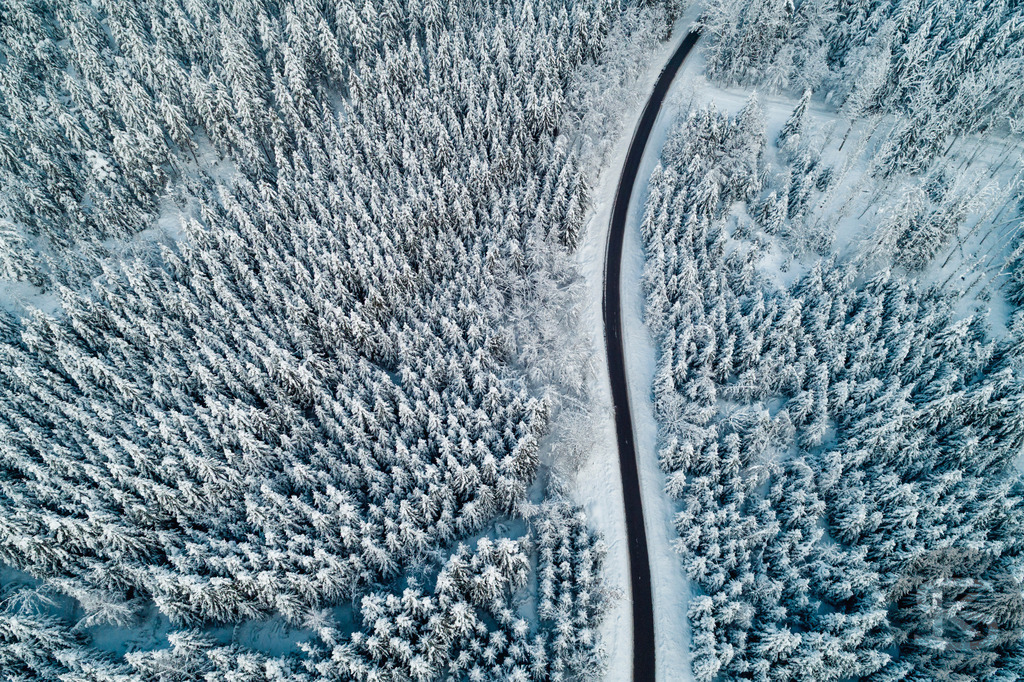 Winterwald aus der Luft – Verschneite Straße durch gefrorenen Nadelwald | Diese Drohnenaufnahme zeigt eine kurvenreiche Straße, die sich durch einen dicht verschneiten Nadelwald schlängelt. Aus der Vogelperspektive wird die geometrische Struktur sichtbar: Die organische Linie der Straße bildet einen starken Kontrast zu den vertikalen Mustern der schneebedeckten Bäume. Die kalten Blautöne verstärken die winterliche Atmosphäre und verleihen dem Wald eine fast abstrakte Qualität. - Realisiert mit Pictrs.com