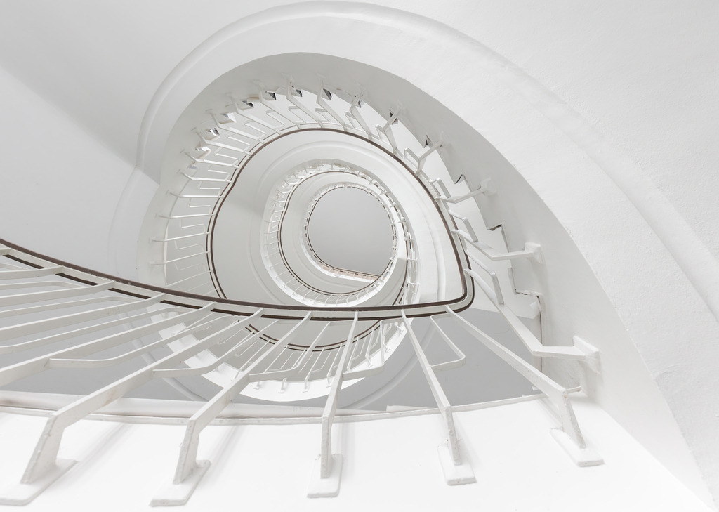 Wandbild Spirale der Unendlichkeit: Blick in ein Treppenhaus | Das Bild zeigt eine beeindruckende spiralförmige Treppe, aufgenommen aus der Perspektive, die nach oben in die Unendlichkeit führt. Die weiße Struktur und die gleichmäßigen, eleganten Geländer schaffen ein faszinierendes geometrisches Muster, das den Blick des Betrachters in die Tiefe der Spirale zieht. Die klare, minimalistische Ästhetik des Treppenhauses betont die Reinheit und Symmetrie des Designs und erzeugt ein Gefühl von Ruhe und Unendlichkeit.
