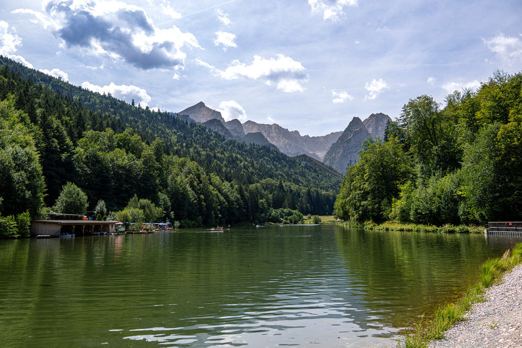 Sommeridylle am Riessersee: Entspannung mit Blick auf die Alpspitze | Das ruhige, grün schimmernde Wasser des Riessersees spiegelt die sanften Wellen eines perfekten Sommertages wider. Umrahmt von dichten, tiefgrünen Mischwäldern öffnet sich das Tal und gibt den Blick frei auf die eindrucksvolle Bergkulisse des Wettersteingebirges mit der markanten Pyramidenspitze der Alpspitze. Am linken Ufer laden Bootssteg und Seebad zum Verweilen ein. Die Aufnahme vermittelt die wohltuende Stille der Natur, die Frische des Wassers und die unbeschwerte Leichtigkeit eines Urlaubs in den bayerischen Alpen. - Realisiert mit Pictrs.com