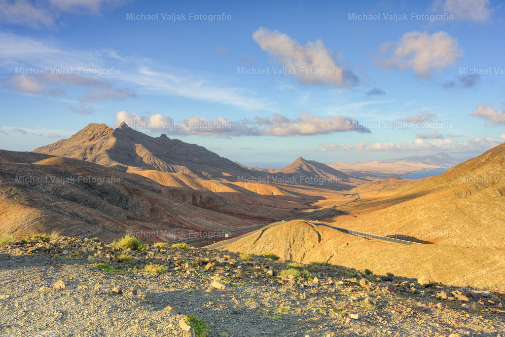 Mirador Astronómico de Sicasumbre auf Fuerteventura | Vom Mirador Astronómico de Sicasumbre öffnet sich der Blick weit über den Süden Fuerteventuras. Die Landschaft fällt in sanften Ebenen ab und führt den Blick direkt auf den Montaña Cardón, der ruhig und markant in der Ferne steht. Das Licht betont die klaren Formen der Hügel und die weiten Flächen dazwischen. Ein Moment, der die stille Größe dieser kargen Vulkanlandschaft spürbar macht. - Realisiert mit Pictrs.com