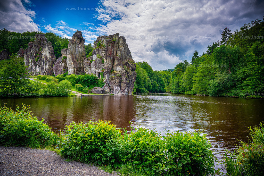 Externsteine im Spiegelbild des Sees | Diese atemberaubende Kunstfotografie fängt die ikonische Felsformation der Externsteine im deutschen Teutoburger Wald ein, die sich dramatisch über einen ruhigen, bernsteinfarbenen See erhebt. Die hoch aufragenden Sandsteinsäulen dominieren die Bildmitte, wobei ihre rauen, verwitterten Oberflächen in warmen, erdigen Rost-, Braun- und Ockertönen leuchten. Ein dichter, üppig grüner Wald umgibt die Felsen zu beiden Seiten und bildet einen natürlichen Rahmen, der den Blick direkt auf die Felsformation lenkt. Darüber verleiht ein stimmungsvoller Himmel voller wogender Wolken der Szene echte Spannung und Tiefe. Das Wasser im Vordergrund spiegelt die Felsen und den Himmel fast perfekt wider und verdoppelt so die visuelle Wirkung. Die Farbgebung tendiert zu satten, intensiven Grüntönen und tiefen, dramatischen Blautönen – was dem gesamten Bild eine filmische, fast jenseitige Atmosphäre verleiht. Es ist die Art von Aufnahme, bei der man innehalten und staunen muss. - Realisiert mit Pictrs.com
