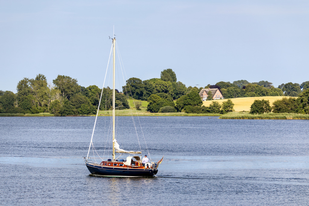Wandbild: Segelboot auf der Schlei in Lindaunis | Dieses Wandbild im Querformat zeigt ein Segelboot auf der Schlei in Lindaunis im Frühling. Das Segelboot spiegelt sich leicht im Wasser der Schlei. Auf der anderen Uferseite sind viele frühlingshafte Bäume und Felder zu sehen. Direkt hinter einem Feld kann man ein Haus erahnen. Der blaue Himmel ist wolkenlos.  - Realisiert mit Pictrs.com