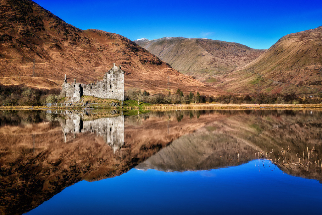 Kilchurn Castle, Loch Awe, Schottland | Die historische Ruine von Kilchurn Castle thront am Ufer von Loch Awe und spiegelt sich perfekt im ruhigen Wasser. Umgeben von den majestätischen, herbstlichen Bergen der schottischen Highlands unter einem klaren blauen Himmel, bietet die Szene eine ikonische Landschaft. Diese Aufnahme betont die dramatische Schönheit und die unberührte Natur der Region. - Realized with Pictrs.com