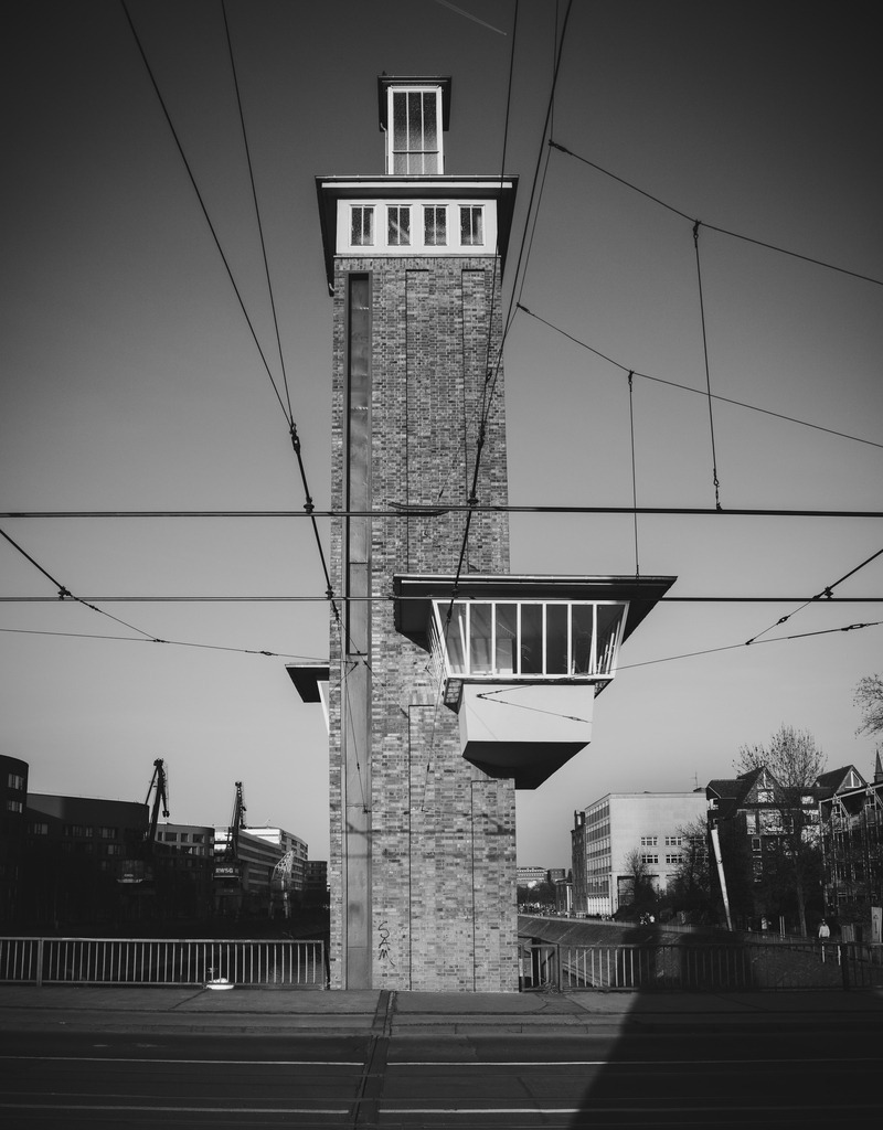  43 Schwanentorbrücke – Duisburger Innenhafen | Eine minimalistische Schwarzweißaufnahme des Schwanentor-Turms im Duisburger Innenhafen, reduziert auf klare Formen, Linien und Struktur. Die markante vertikale Architektur wird von einem feinen Netz aus Oberleitungen durchzogen, das die Komposition aufbricht und zugleich eine spannende grafische Spannung erzeugt.Das Zusammenspiel aus strenger Geometrie und filigranen Linien verleiht dem Motiv eine präzise, fast technische Ästhetik. In monochromer Darstellung tritt die Architektur in den Vordergrund und entwickelt eine ruhige, zugleich kraftvolle Wirkung – geprägt von Klarheit, Struktur und urbaner Präsenz.Diese Aufnahme übersetzt die urbane Architektur des Ruhrgebiets in eine zeitlose Fine-Art-Komposition, die sich besonders für moderne, designorientierte Räume eignet. - Realisiert mit Pictrs.com