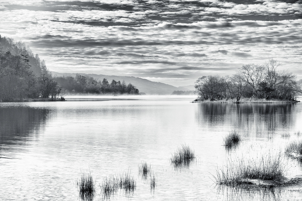 Loch Awe: Winterliche Morgenlandschaft | Eine Schwarz-Weiss-Aufnahme fängt die ruhige Weite von Loch Awe in Schottland ein, mit einer bewaldeten Insel im Vordergrund und nebelverhangenen Bergen im Hintergrund. Die Komposition betont die Texturen des Himmels, die Spiegelungen auf dem Wasser und die kahlen Bäume der winterlichen Landschaft, eingefangen an einem Wintermorgen. - Realisiert mit Pictrs.com