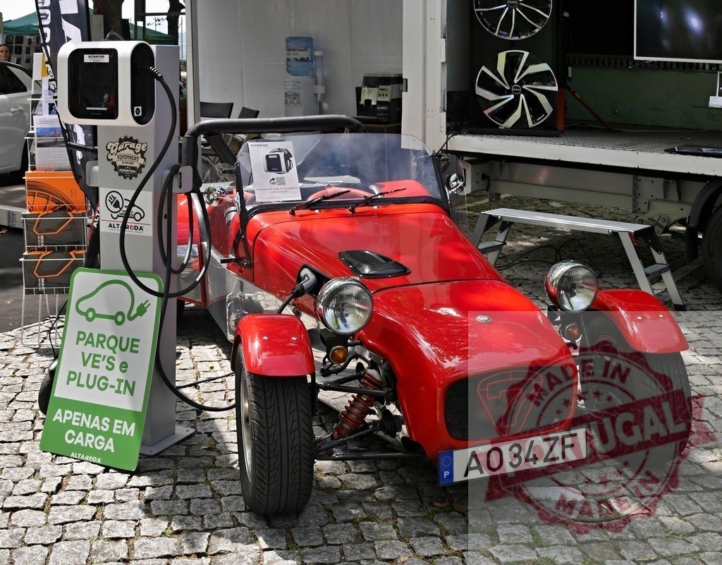 Red electric Caterham 7 roadster charging at station | Red Caterham 7 electric car plugging in at outdoor charging station - Realized with Pictrs.com