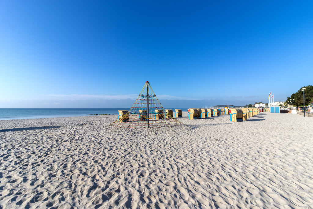 Wandbild: Kletterspaß am Meer – Grömitz mit Pyramide | Dieses Wandbild zeigt eine lebendige Szene vom Strand in Grömitz. Im Zentrum des Bildes steht eine Kletterpyramide aus Seilen – ein beliebtes Spielgerät für Kinder, das Bewegung und Abenteuer direkt am Meer ermöglicht. Rund um die Pyramide sind zahlreiche Strandkörbe in geordneter Reihe aufgestellt, alle ausgerichtet zum Wasser hin. Der Strand ist weitläufig und gepflegt, der Himmel klar und das Meer ruhig – perfekte Bedingungen für einen entspannten Sommertag. Die Komposition verbindet Freizeitspaß mit maritimer Gelassenheit und eignet sich ideal als Wandbild für familienfreundliche Wohnkonzepte oder Ferienunterkünfte. Erhältlich als Leinwandbild, Acrylglasbild, Alu-Dibond FineArt Print oder als Akustikbild. - Realisiert mit Pictrs.com