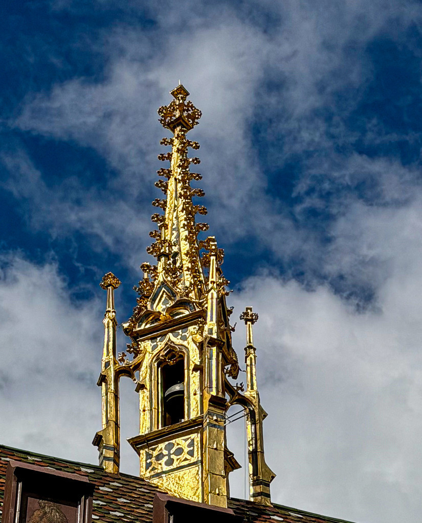 Basel Rathaus – Majestätische goldene Turmspitze Architektur Detail | Exklusive Architekturfotografie aus Basel: Dieses High-End Wandbild zeigt die prunkvolle, goldene Spitze des historischen Basler Rathauses. Vor einem dramatischen, tiefblauen Wolkenhimmel entfaltet das filigrane goldene Meisterwerk zusammen mit den traditionell gemusterten Basler Dachziegeln seine volle visuelle Pracht. Ein charakterstarkes, elegantes Kunstwerk in Galeriequalität – der perfekte Blickfang für anspruchsvolle Wohnräume und Büros. - Realisiert mit Pictrs.com