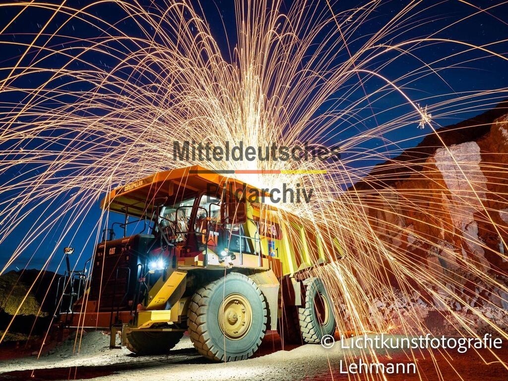 Lichtkunst_Mammendorf_Kipper_7674 | Das Foto zeigt einen Muldenkipper vom Typ Caterpillar CAT 775G im Steinbruch Mammendorf aus einer bodennahen Perspektive. Die gewaltige Baumaschine steht im Mittelpunkt der Aufnahme und verdeutlicht eindrucksvoll die Dimensionen moderner Gewinnungstechnik im Gesteinsabbau. Durch die tiefe Kameraposition wirkt der Muldenkipper besonders imposant und unterstreicht seine enorme Leistungsfähigkeit.Der CAT 775G wird von einem 16-Zylinder-Dieselmotor mit 765 PS (563 kW) angetrieben. Die Maschine besitzt eine Nutzlast von bis zu 64 Tonnen und erreicht ein Gesamtgewicht von rund 125 Tonnen. Mit einer Höchstgeschwindigkeit von etwa 67 km/h transportiert der Großmuldenkipper täglich tausende Tonnen Gestein zwischen Abbauwand und Brechanlage und zählt zu den leistungsstärksten Transportfahrzeugen seiner Klasse im Steinbruchbetrieb.Die Aufnahme entstand als Langzeitbelichtung des Lichtkunstfotografen Daniel Lehmann. Während der Belichtungszeit wurden Lichteffekte und Feuer gezielt eingesetzt, um die kraftvolle Technik des Muldenkippers künstlerisch in Szene zu setzen. Das Zusammenspiel aus moderner Schwerlasttechnik, Lichtkunst und Fotografie verleiht dem Bild eine außergewöhnliche Dynamik und macht die Leistungsfähigkeit dieser imposanten Arbeitsmaschine eindrucksvoll erlebbar. - Realisiert mit Pictrs.com